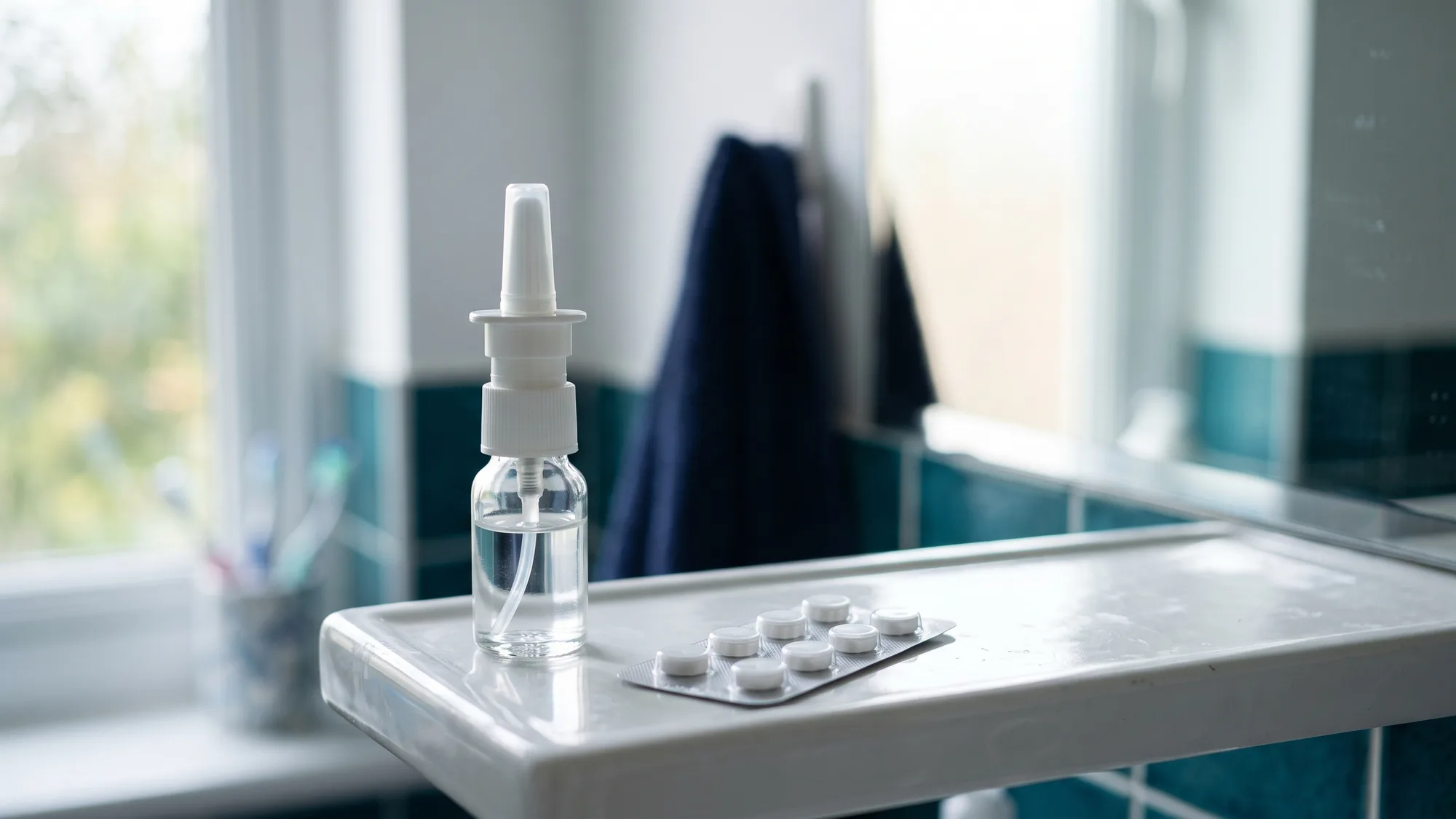 Opakowanie leku przeciwbólowego i mały spray do nosa leżą na białej półce łazienkowej, w tle rozmyte lustro i poranne światło z okna